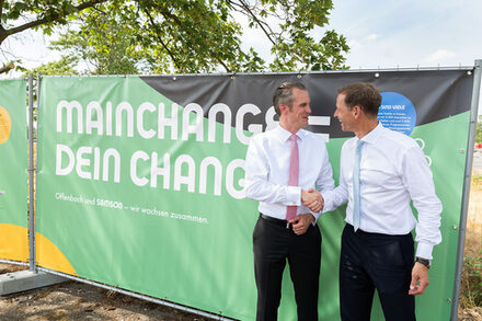 Zwei Männer geben sich vor einem Bauzaun, auf dem „MainChance = Dein Change – Offenbach und Samson wachsen zusammen“ steht, die Hände.