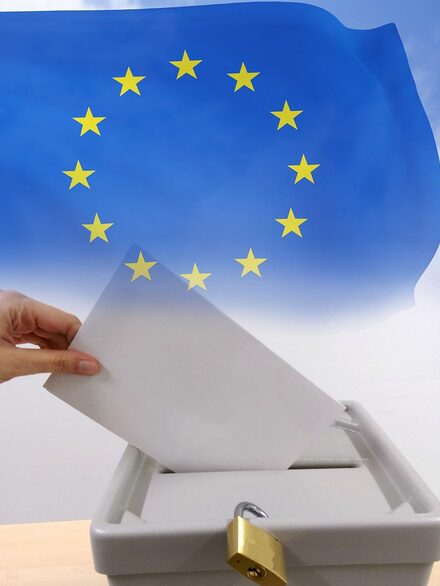 Eine Hand wirft einen Wahlbrief in eine Wahlurne. Dahinter ist die Europafahne zu sehen.
