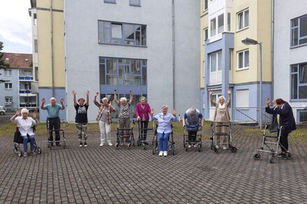 Seniorinnen und Senioren stehen mit ihren Rollatoren auf einem Platz und heben die Arme.