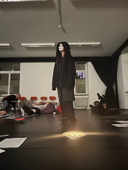 Eine Figur mit Maske steht im Raum, andere Menschen liegen drumherum.