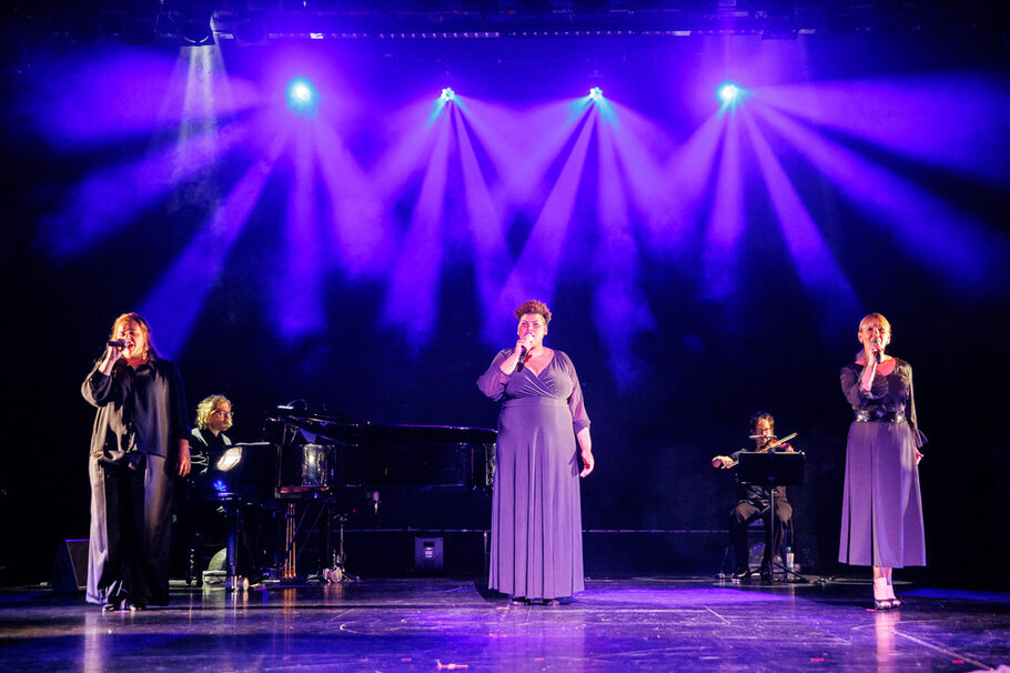Drei Sängerinnen und zwei Musiker auf einer lila beleuchteten Bühne