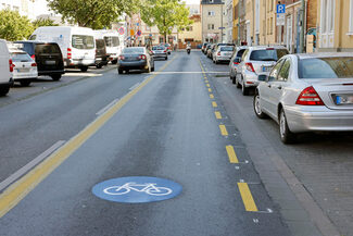 Auf dem Foto ist eine Straße, parkende Auto und eine Fahrradspur zu sehen.