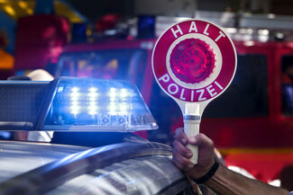 Eine Hand hält eine Kelle mit der Aufschrift "Halt Polizei" neben einem Blaulicht eines Polizeiautos.