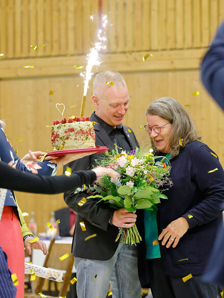 Ein Mann und eine Frau halten einen Blumenstrauß und freuen sich, im Vordergrund eine Torte mit Minifeuerwerk.