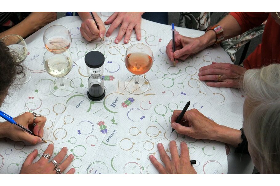 Das Foto zeigt einen Tisch mit Weingläsern und Vordrucken für ein Textspiel. Die Vordrucke werden von mehreren Personen ausgefüllt.