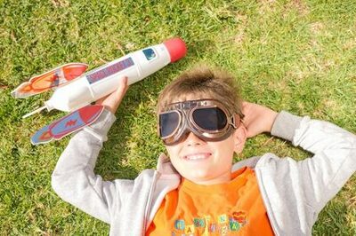 Kind mit einer Fliegerbrille und einer Rakete in der Hand liegt auf einer Wiese